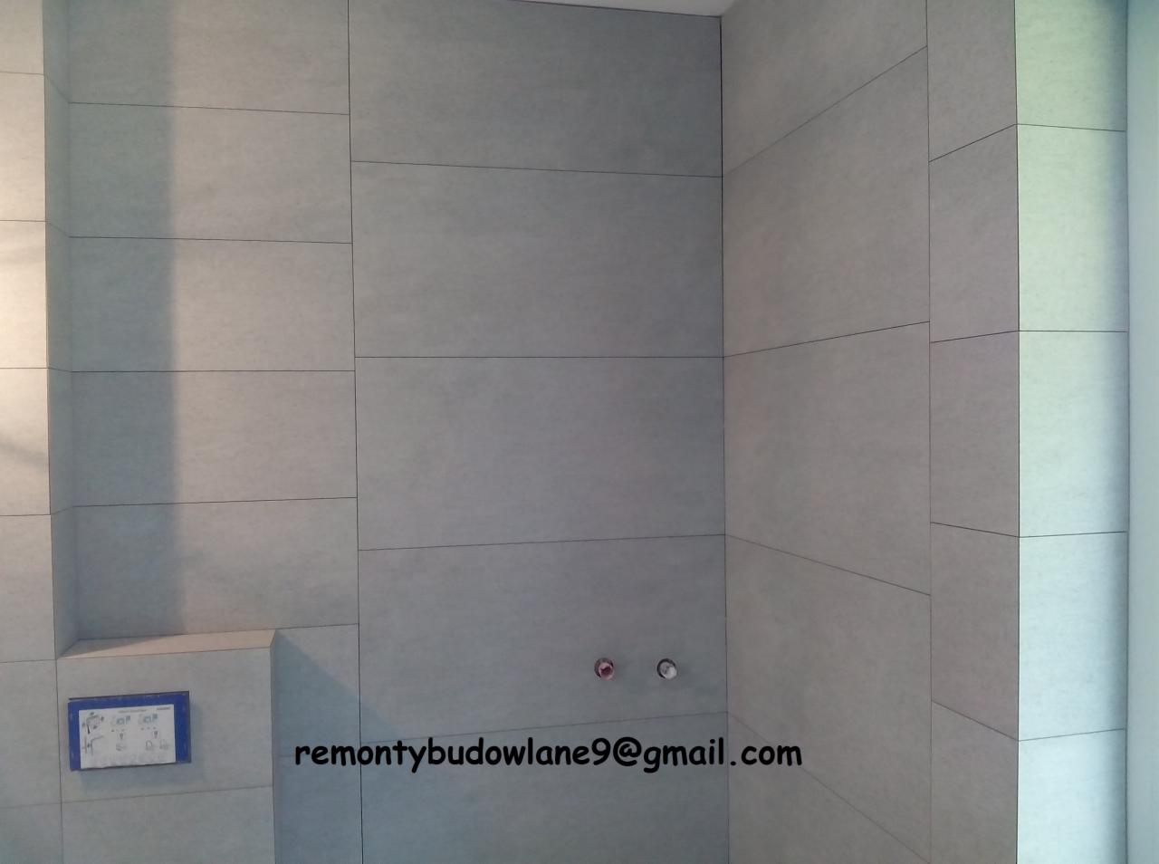 Wykończona ściana łazienki z szarymi, prostokątnymi płytkami ceramicznymi, widoczne przyłącza wodne i podtynkowy stelaż WC z niebieską ramką.