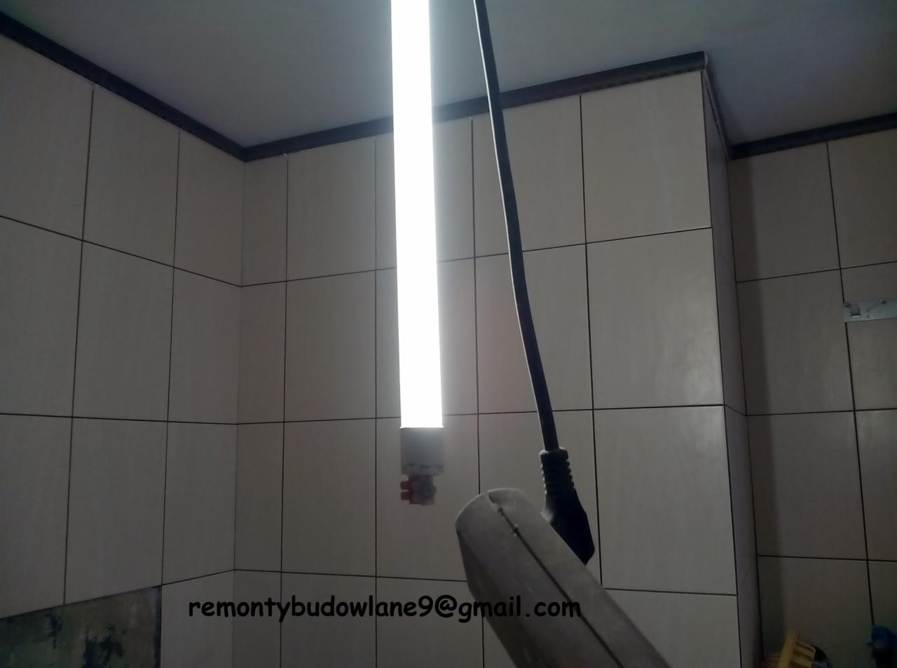 Wykończona ściana w łazience z jasnymi, kwadratowymi płytkami ceramicznymi i ciemną fugą, oświetlona pionową lampą roboczą. W kadrze fragment narzędzia. Na dole widoczny adres email.