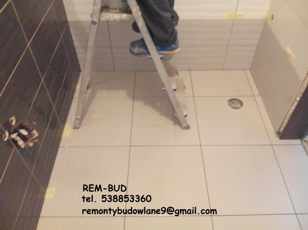 Wnętrze łazienki w trakcie układania płytek, widoczny fragment ściany z brązowymi płytkami, białe płytki na podłodze i ścianach, drabina aluminiowa z pracownikiem, taśma malarska zabezpieczająca...