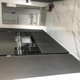 Prestige - Częściowo wykończona kuchnia z ciemnymi, matowymi szafkami i białymi płytkami imitującymi marmur na podłodze. W tle widoczny niezabudowany fragment ściany, sugerujący trwający remont.