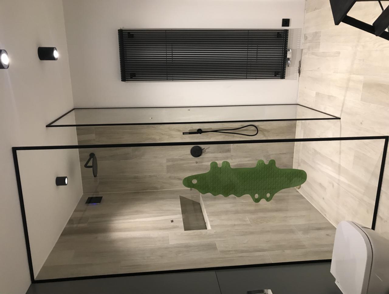Nowoczesna kabina prysznicowa typu walk-in z czarnymi profilami, wyłożona płytkami imitującymi drewno, z zieloną wycieraczką w kształcie krokodyla i minimalistycznymi lampami punktowymi.