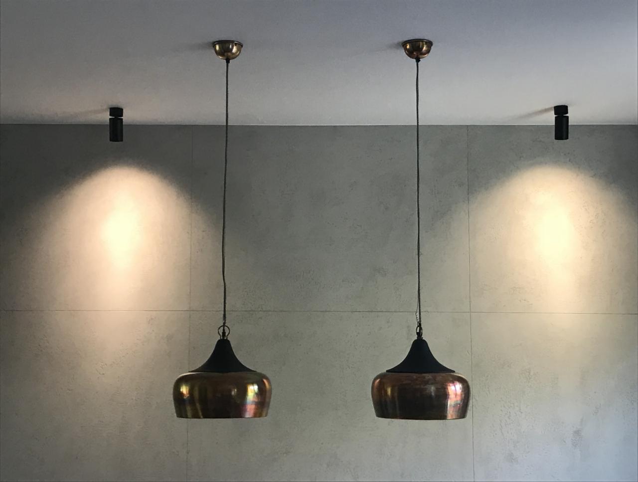 Dwa miedziane klosze lamp wiszących z czarnymi elementami, zawieszone na tle szarej ściany z widocznymi łączeniami płyt i punktowymi reflektorami sufitowymi.
