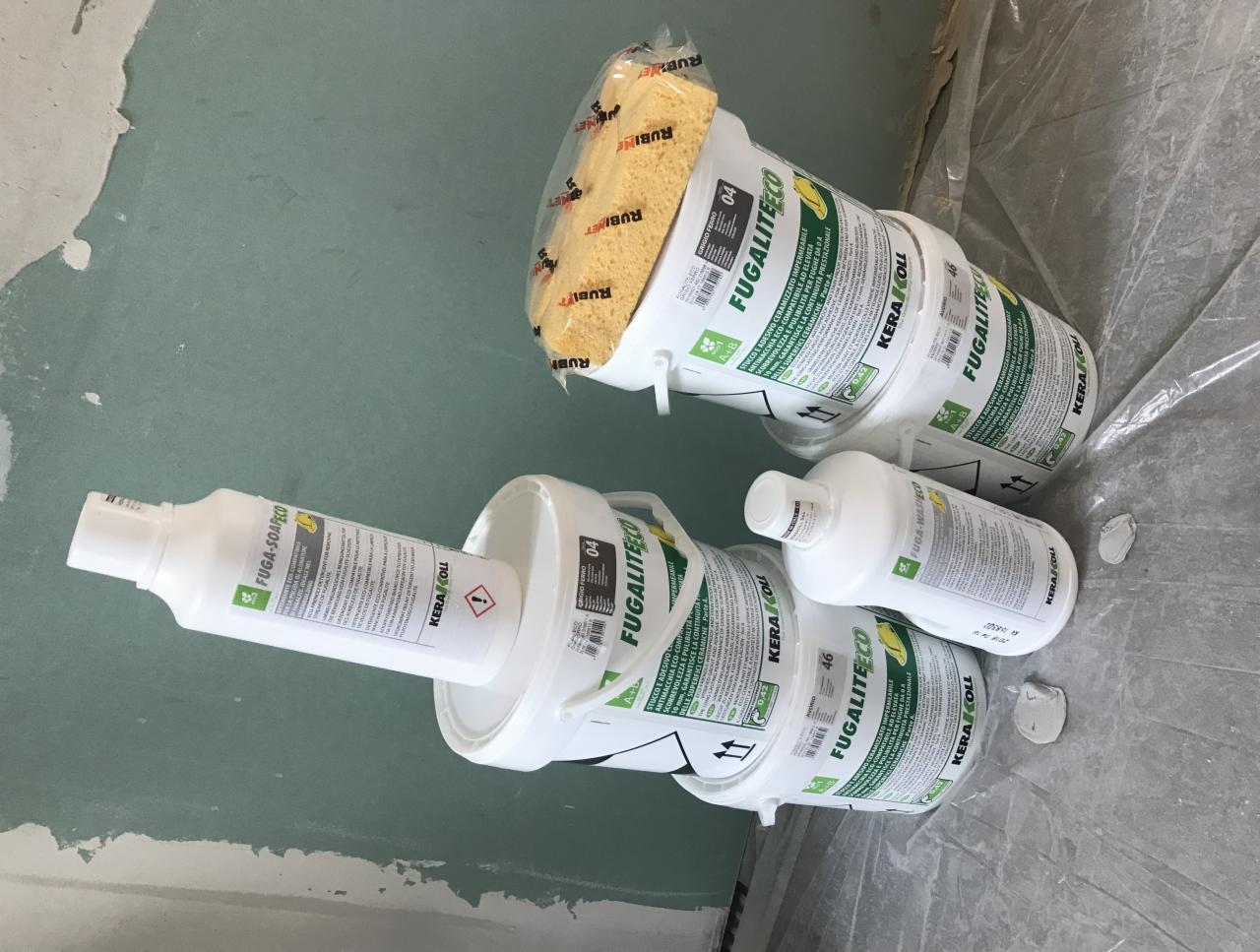 Pakiety i pojemniki z materiałami do fugowania i czyszczenia płytek, widoczne wiadra z Fugalite Eco, gąbka Rubi i butelka Fuga-Soap Eco na podłodze w trakcie prac wykończeniowych.