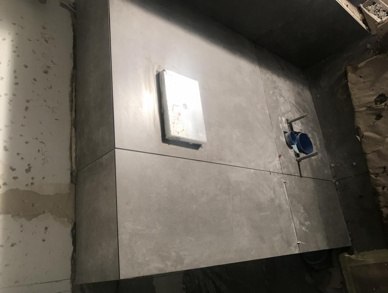 Szare, wielkoformatowe płytki ścienne w trakcie montażu w łazience, widoczne krzyżyki dystansowe, nieotynkowana ściana z lewej strony i instalacja podtynkowa.