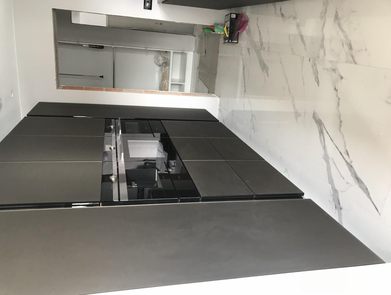 Częściowo wykończona kuchnia z ciemnymi, matowymi szafkami i białymi płytkami imitującymi marmur na podłodze. W tle widoczny niezabudowany fragment ściany, sugerujący trwający remont.