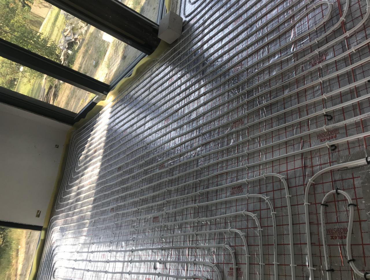 Montaż ogrzewania podłogowego w pomieszczeniu z dużym oknem w Warszawie, widoczne rury grzewcze ułożone na izolacji z siatką.