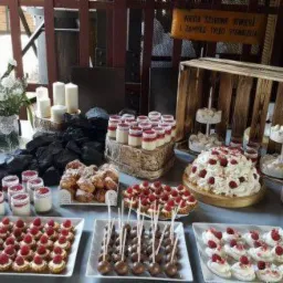 Stół z deserami na przyjęciu w Grodzisku Wielkopolskim: tort bezowy z malinami, babeczki, mini desery w słoiczkach, pączki i trufle czekoladowe, dekoracje z węgla i świec.