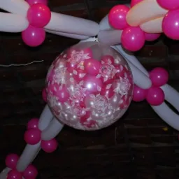 Dekoracja balonowa: duży przezroczysty balon z różowymi balonikami wewnątrz, ozdobiony białymi wzorami róż, otoczony girlandą z białych i różowych balonów.