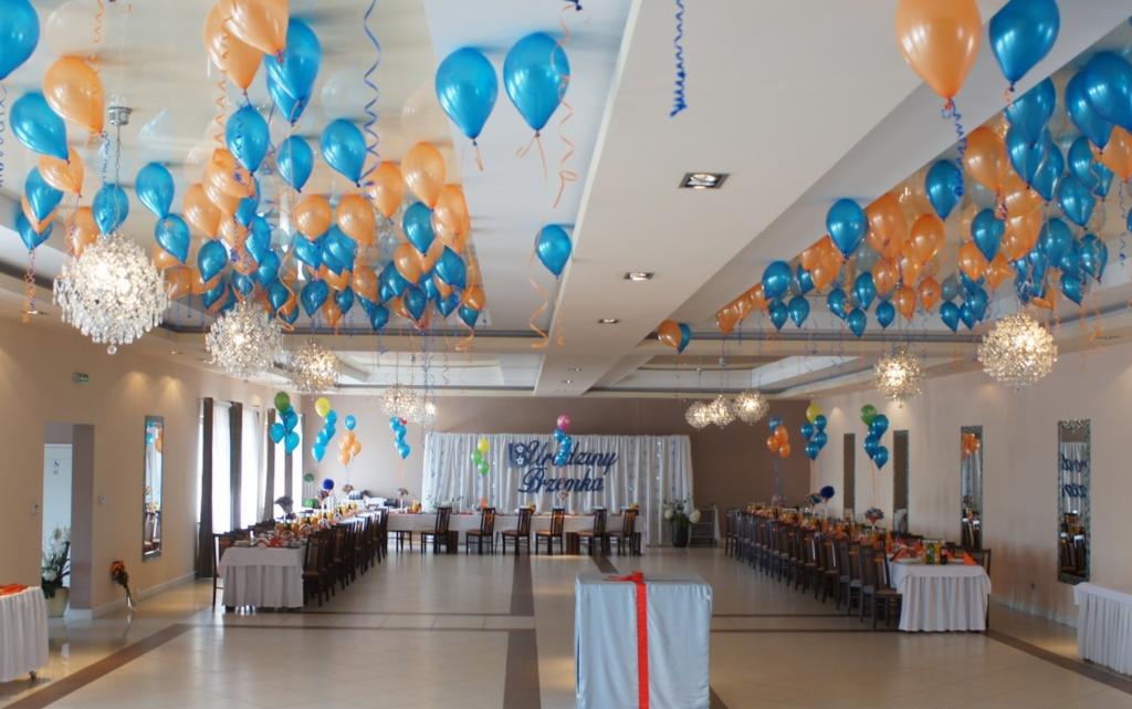 Udekorowana sala balowa z niebieskimi i pomarańczowymi balonami pod sufitem, kryształowe żyrandole, stoły nakryte białymi obrusami, scena z napisem 'Uroczy Dzień', duży prezent na pierwszym planie.