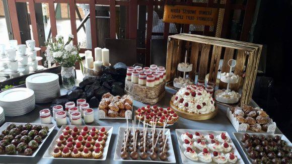 Stół z deserami na przyjęciu w Grodzisku Wielkopolskim: tort bezowy z malinami, babeczki, mini desery w słoiczkach, pączki i trufle czekoladowe, dekoracje z węgla i świec.