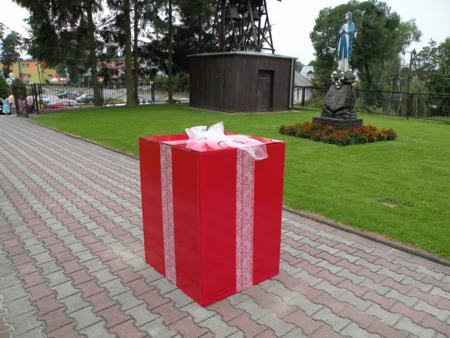 Duży, czerwony prezent z białą kokardą na chodniku, w tle drewniana budowla i figura religijna.