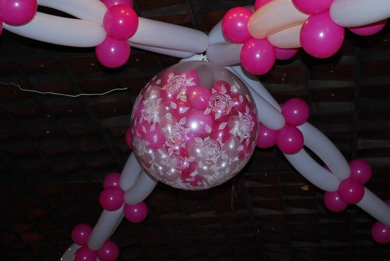 Dekoracja balonowa: duży przezroczysty balon z różowymi balonikami wewnątrz, ozdobiony białymi wzorami róż, otoczony girlandą z białych i różowych balonów.