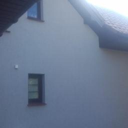 macedonska - Elewacja domu jednorodzinnego w odcieniach szarości z dwoma oknami o ciemnych ramach, widoczne fragmenty dachu z brązową dachówką i ciemną podbitką, oświetlone słońcem.