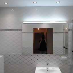 macedonska - Wykończona łazienka z białą umywalką i lustrem, na ścianach białe płytki z delikatnym wzorem rombów i szarym paskiem dekoracyjnym, widoczne oświetlenie sufitowe i nad lustrem, odbicie osoby...