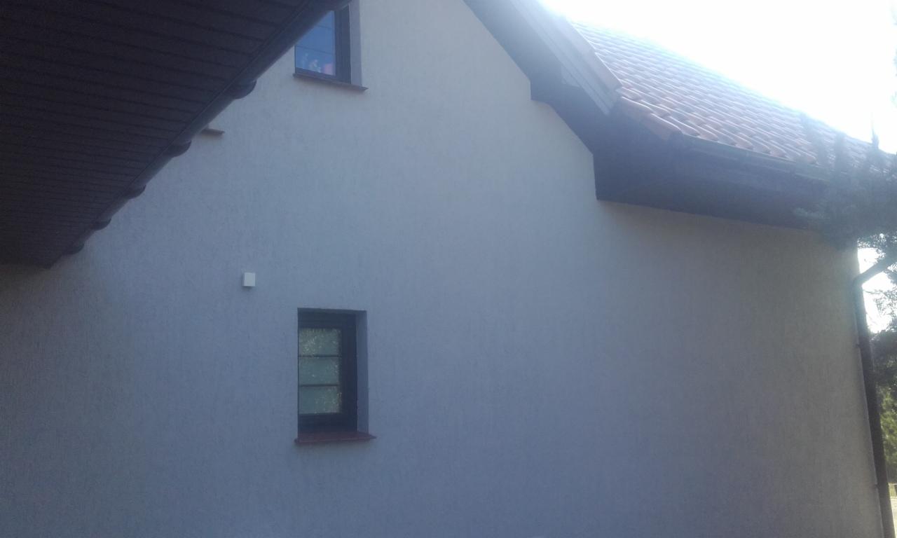 Elewacja domu jednorodzinnego w odcieniach szarości z dwoma oknami o ciemnych ramach, widoczne fragmenty dachu z brązową dachówką i ciemną podbitką, oświetlone słońcem.