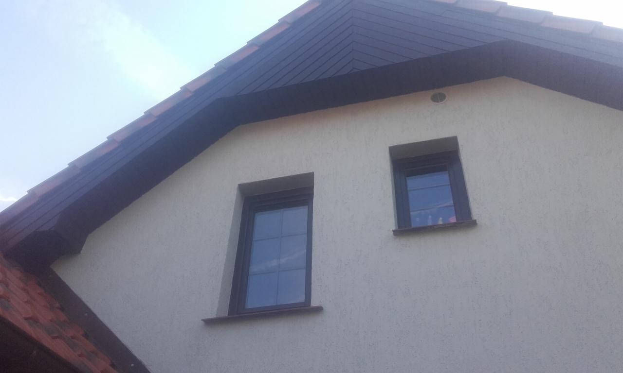 Fragment elewacji domu z dwoma oknami o różnej wielkości i ciemnobrązowym dachem z wystającym okapem.