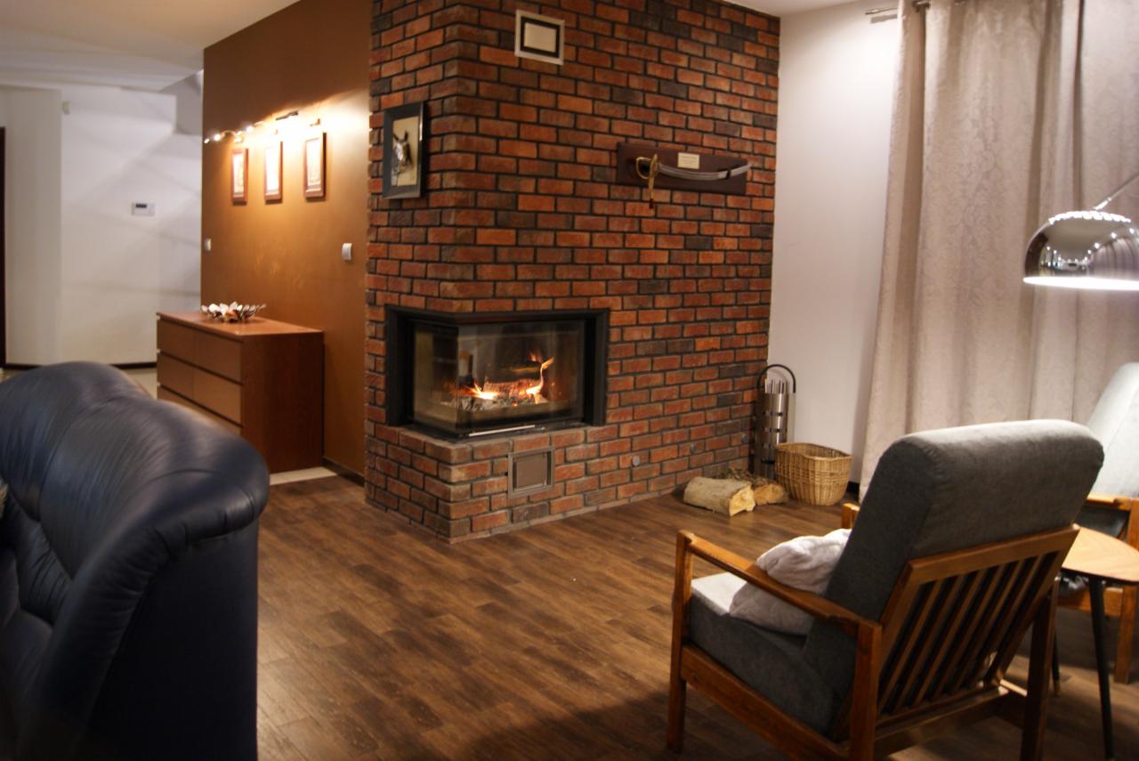 Salon z ceglanym kominkiem narożnym, płomieniami w palenisku, drewnianą podłogą, szarym fotelem i elementami dekoracyjnymi na ścianie.