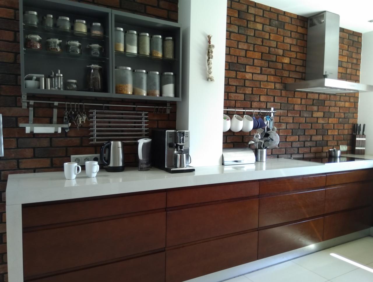 Nowoczesna kuchnia z ceglaną ścianą, drewnianymi szafkami, białym blatem, szklanymi pojemnikami na przyprawy i wiszącym czosnkiem.