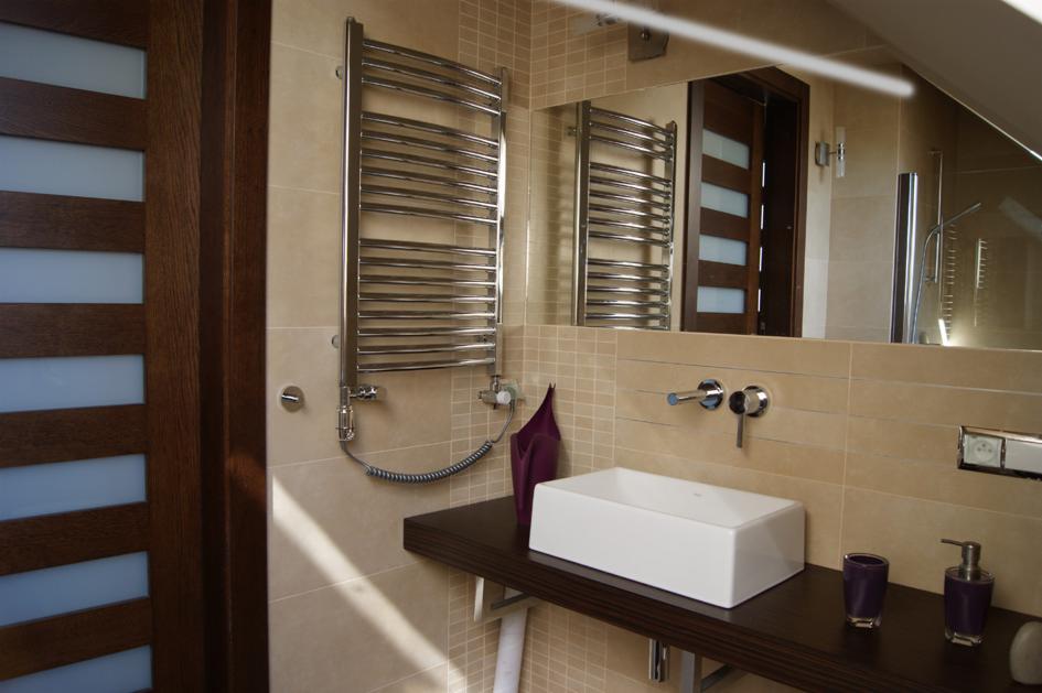 Nowoczesna łazienka z beżowymi płytkami, prostokątną umywalką nablatową, grzejnikiem drabinkowym i drewnianymi drzwiami z mlecznymi szybami w tle.