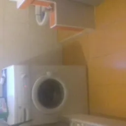 Pralka w łazience z beżowymi i żółtymi płytkami, widoczna półka i okno z detergentami, zdjęcie o słabej jakości.