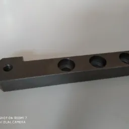 Metalowy element z czterema otworami i gwintem, leżący na białym tle, sfotografowany z bliska.