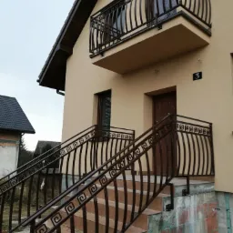 Beżowy dom jednorodzinny z balkonem i schodami wejściowymi, ozdobne metalowe balustrady w kolorze brązowym z motywem greckim, widok z dołu na fragment budynku.