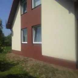 Dwukolorowa elewacja domu jednorodzinnego, z brązowym pasem przy ziemi i wokół okien, oraz beżową resztą ściany, widok z boku na tle trawnika.