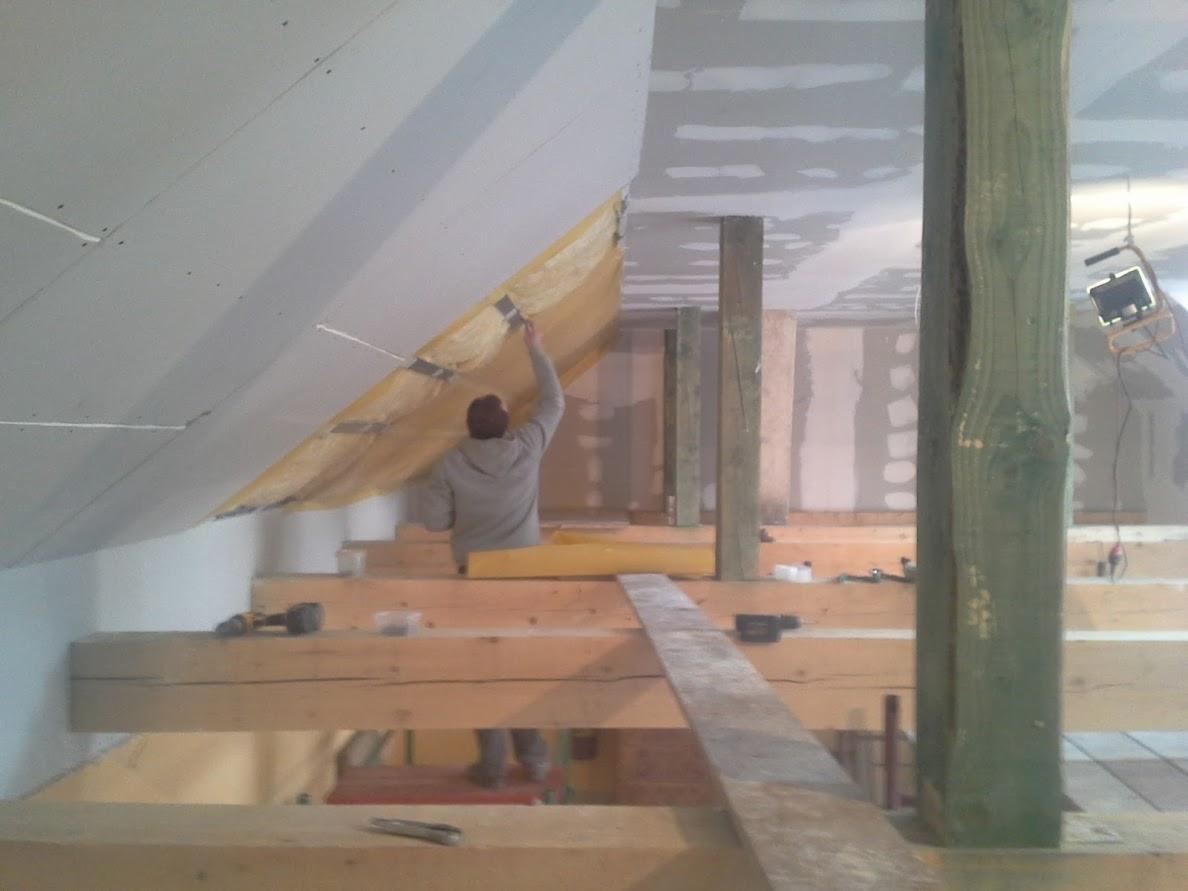 Pracownik mocuje wełnę mineralną do skośnej ściany poddasza, przygotowując ją do montażu płyt kartonowo-gipsowych; widoczne drewniane rusztowania i słupy konstrukcyjne.