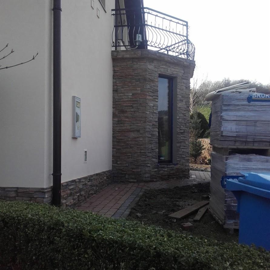 Wykończona elewacja budynku z elementami kamiennymi i balkonem z ozdobną balustradą, widoczne materiały budowlane w tle.