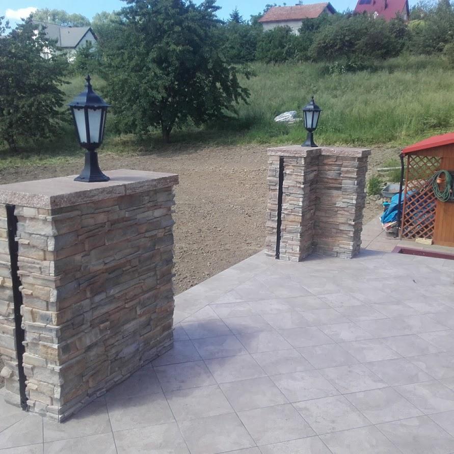 Dwa kamienne słupki z lampami na tle wzgórza i domu, wykończone płytkami tarasowymi w odcieniach szarości.