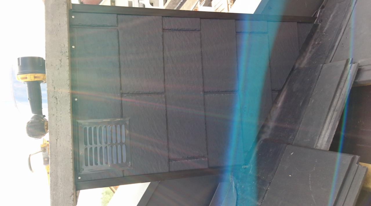 Fragment dachu z zamontowaną wentylacją, pokryty płytami imitującymi łupek, widoczny wiertarko-wkrętak przy betonowym elemencie konstrukcyjnym, silne refleksy świetlne.