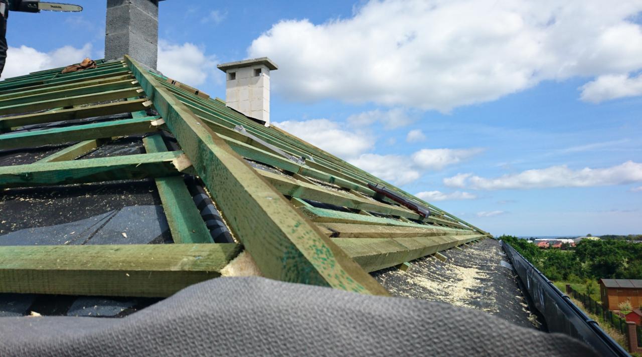 Konstrukcja więźby dachowej z widocznymi elementami drewnianymi i kominem, na tle błękitnego nieba z chmurami.