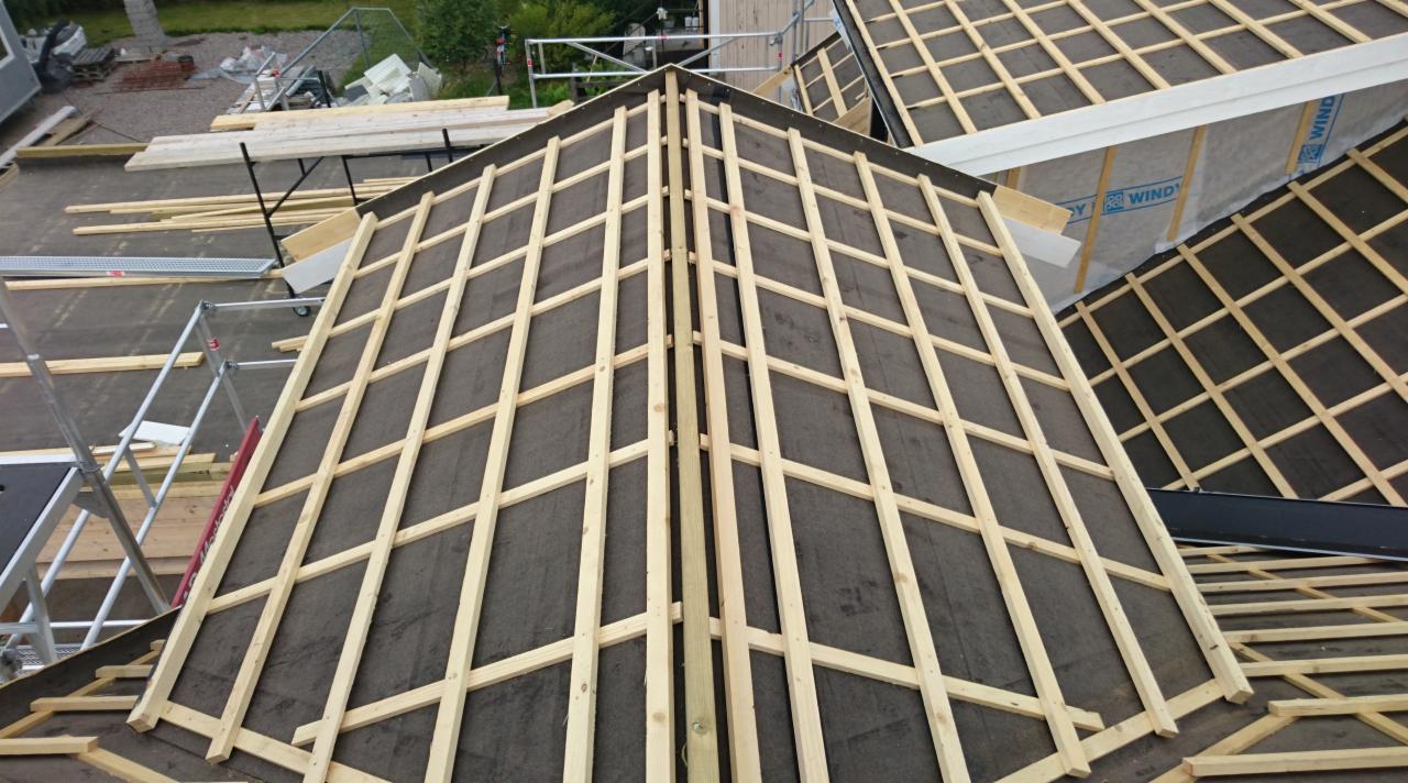 Wiązar dachowy z łatami na czarnej membranie, widok z góry na konstrukcję dachu w trakcie budowy, w tle rusztowania i materiały budowlane.
