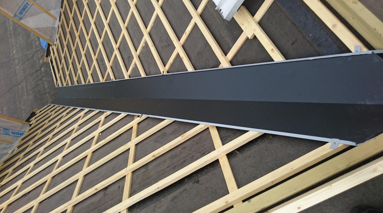 Widok z góry na konstrukcję więźby dachowej z zamontowaną, długą, czarną obróbką blacharską, prawdopodobnie okapową. Widoczna membrana dachowa pod konstrukcją.