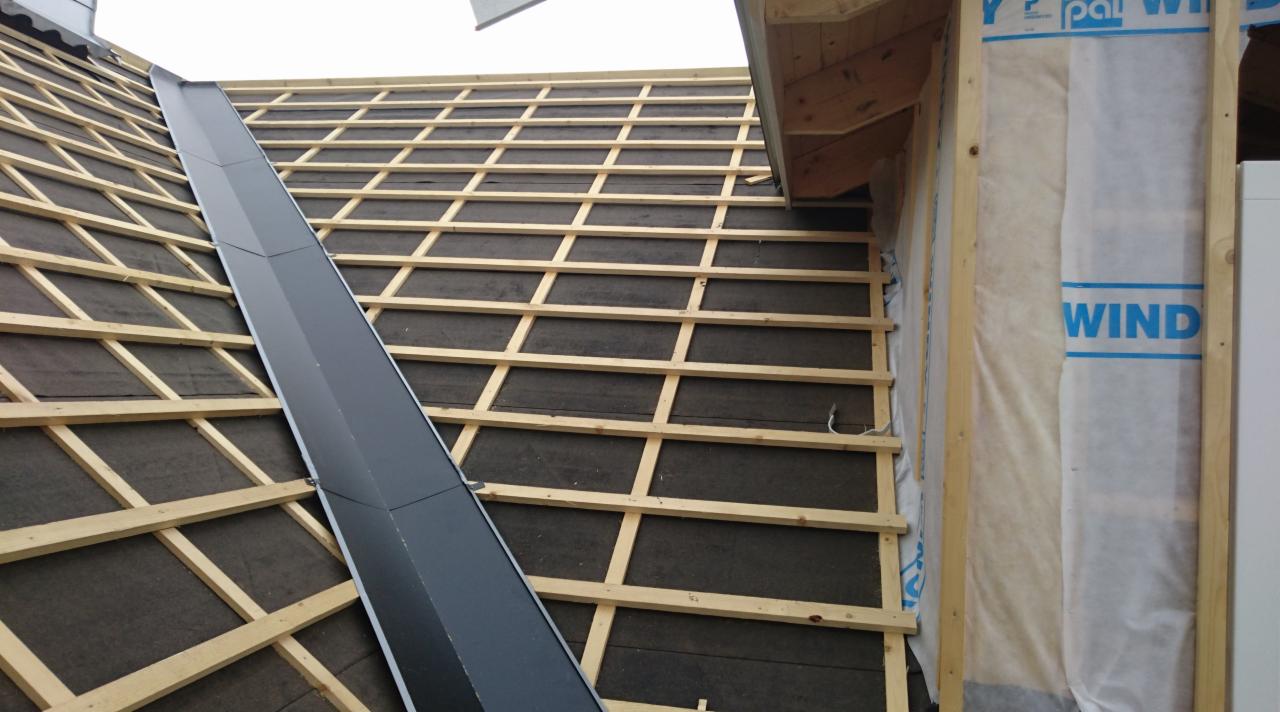 Konstrukcja dachu z widoczną więźbą, czarną membraną i elementami obróbki blacharskiej, z bliska widoczna ściana szkieletowa z izolacją przeciwwiatrową.