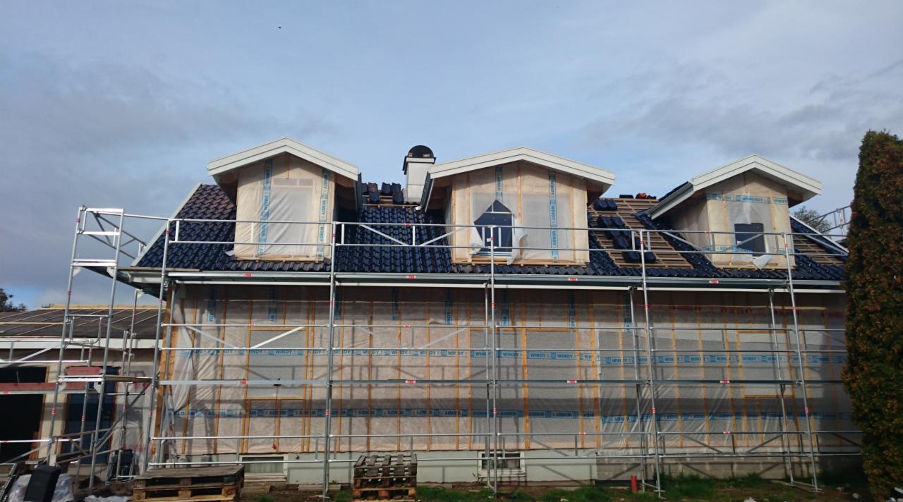 Dom w trakcie budowy z widocznymi rusztowaniami, trzema lukarnami z oknami zabezpieczonymi folią, częściowo ułożonym ciemnym dachem z dachówki oraz kominem.