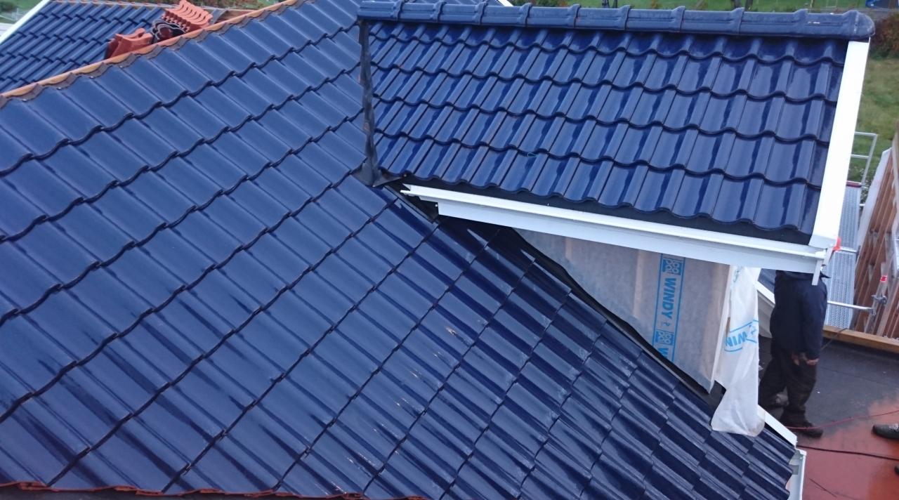 Połacie dachu kryte błyszczącą, granatową dachówką ceramiczną, widoczne obróbki blacharskie, fragment rusztowania i pracownik w tle.
