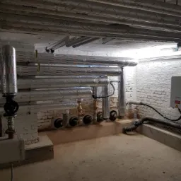 Piwnica z rurami centralnego ogrzewania i instalacji wodnej, pokrytymi izolacją Rockwool, przy ścianie z białej cegły, widoczne zawory i elementy przyłączeniowe.