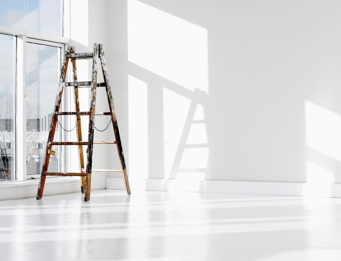 Jasne, minimalistyczne wnętrze z drewnianą drabiną malarską opartą o okno, ślady farby na drabinie, odbicia światła na podłodze.