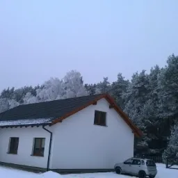 Biały dom jednorodzinny z ciemnym dachem i drewnianą podbitką w zimowej scenerii, flaga Polski na pierwszym planie, za domem ośnieżony las i zaparkowany szary samochód.