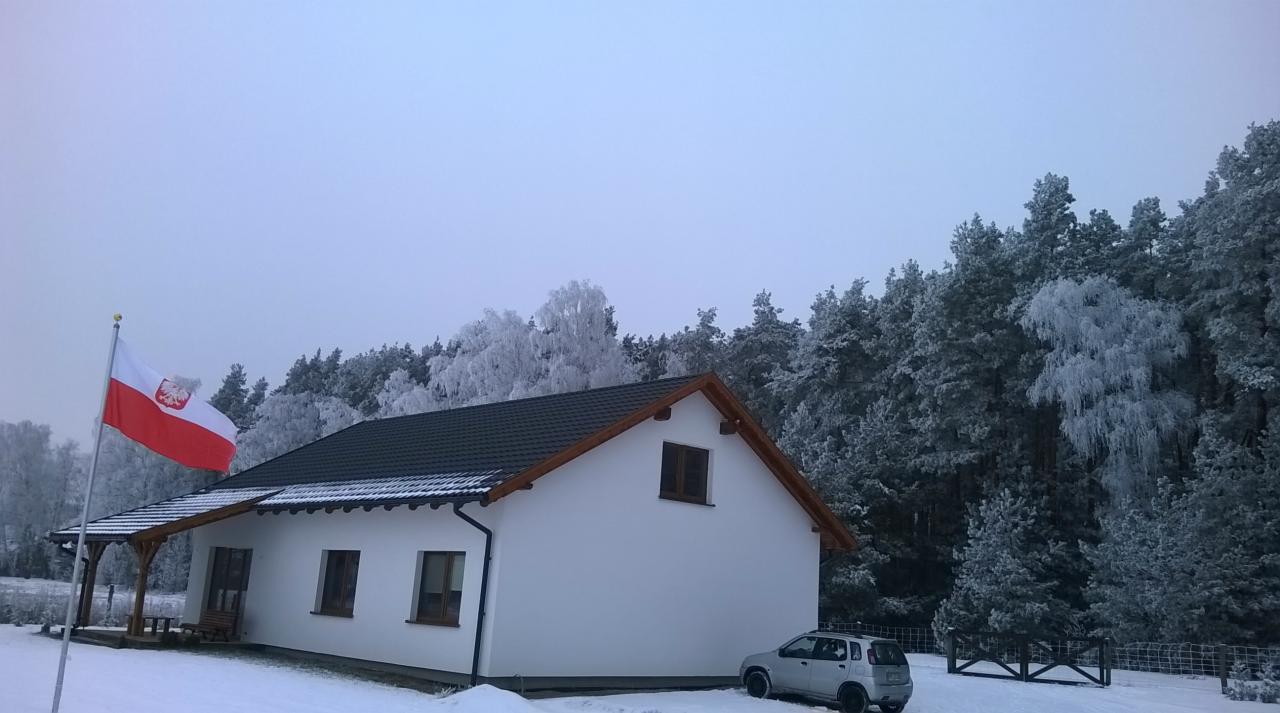 Biały dom jednorodzinny z ciemnym dachem i drewnianą podbitką w zimowej scenerii, flaga Polski na pierwszym planie, za domem ośnieżony las i zaparkowany szary samochód.