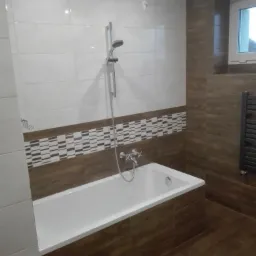 Wykończona łazienka z wanną obudowaną brązowymi płytkami imitującymi drewno, białe płytki na ścianach z dekoracyjnym pasem mozaiki, chromowana bateria wannowa i słuchawka prysznicowa.