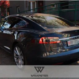 Ciemnoszara Tesla Model S zaparkowana na zewnątrz budynku, widok z tyłu, z delikatnie przyciemnionymi szybami i logo firmy Wrapster na dole zdjęcia.