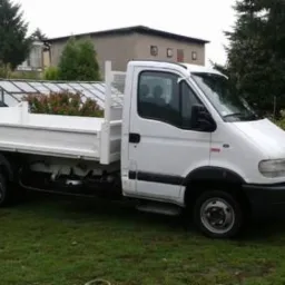 Biały samochód dostawczy z otwartą skrzynią ładunkową, zaparkowany na trawie przed budynkiem mieszkalnym i szklarnią.