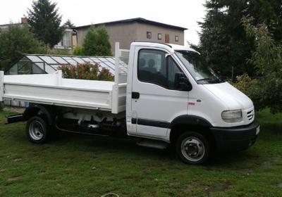 Biały samochód dostawczy z otwartą skrzynią ładunkową, zaparkowany na trawie przed budynkiem mieszkalnym i szklarnią.