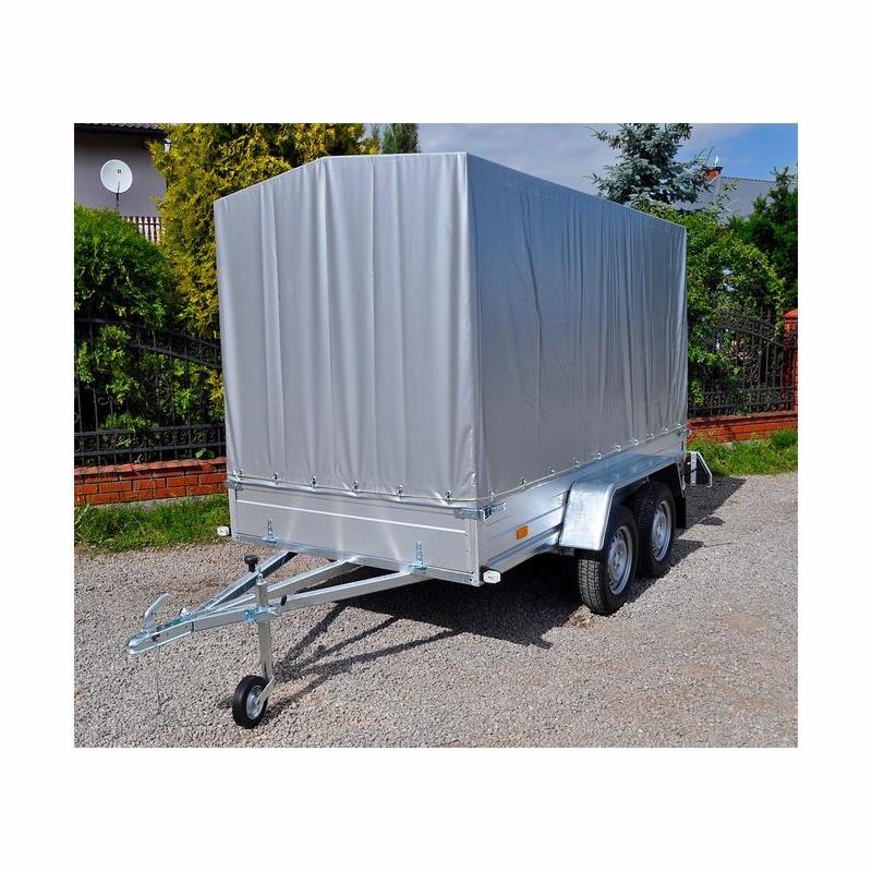 Przyczepa dwuosiowa z plandeką w kolorze srebrnym, zaparkowana na żwirowym podłożu, widok z przodu na tle zieleni.