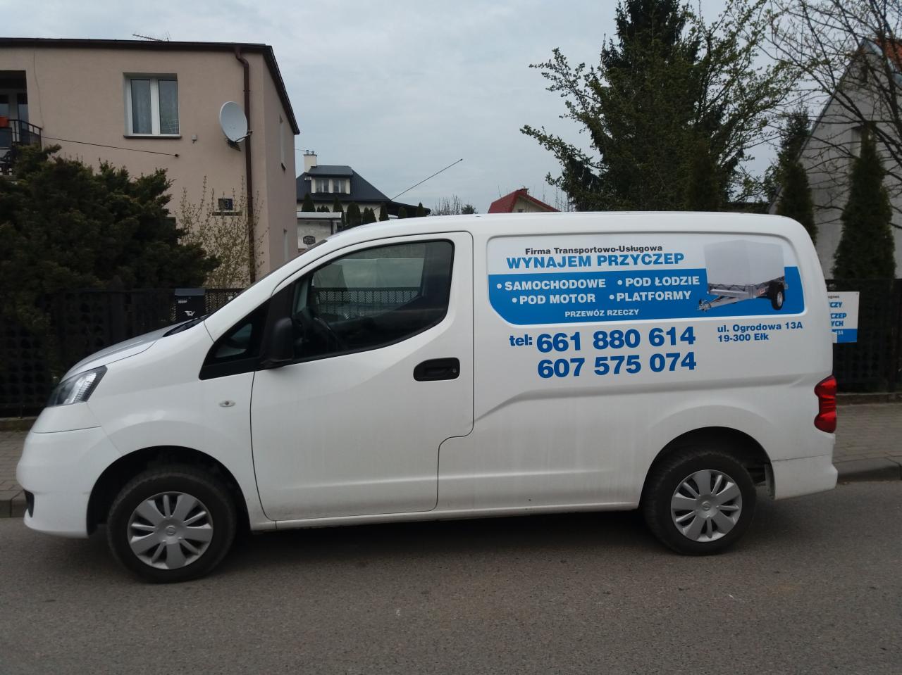 Biały bus firmy transportowej z niebieskim nadrukiem reklamowym, widok z boku na tle budynku mieszkalnego i drzew.