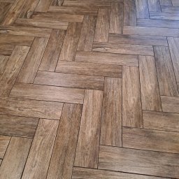 MM Pulut - Ułożona w jodełkę podłoga z płytek imitujących drewno, ciepłe odcienie brązu, widok z góry, fuga w kolorze zbliżonym do płytek, fragment mebla w tle.