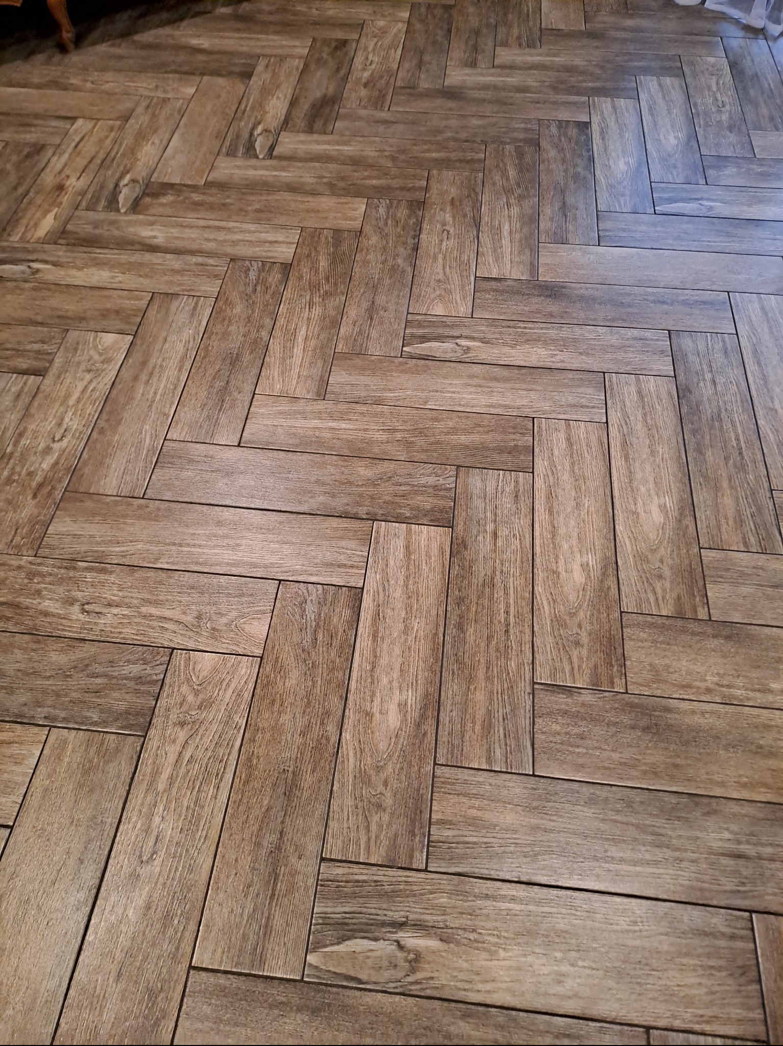 Ułożona w jodełkę podłoga z płytek imitujących drewno, ciepłe odcienie brązu, widok z góry, fuga w kolorze zbliżonym do płytek, fragment mebla w tle.