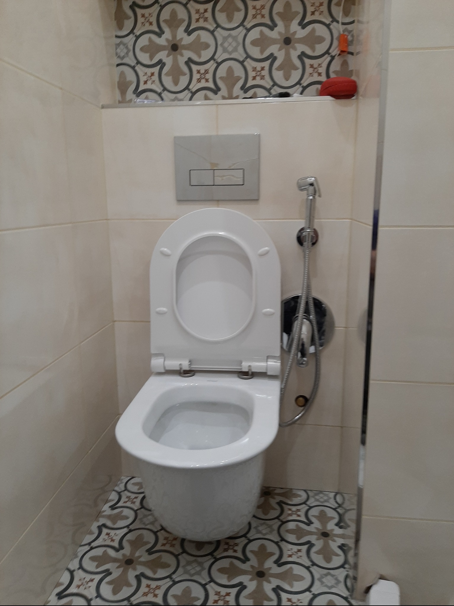 Biała toaleta wisząca z bidetką w wykończonej łazience. Beżowe płytki ścienne i podłogowe z wzorem. Widoczny przycisk spłuczki i akcesoria łazienkowe.